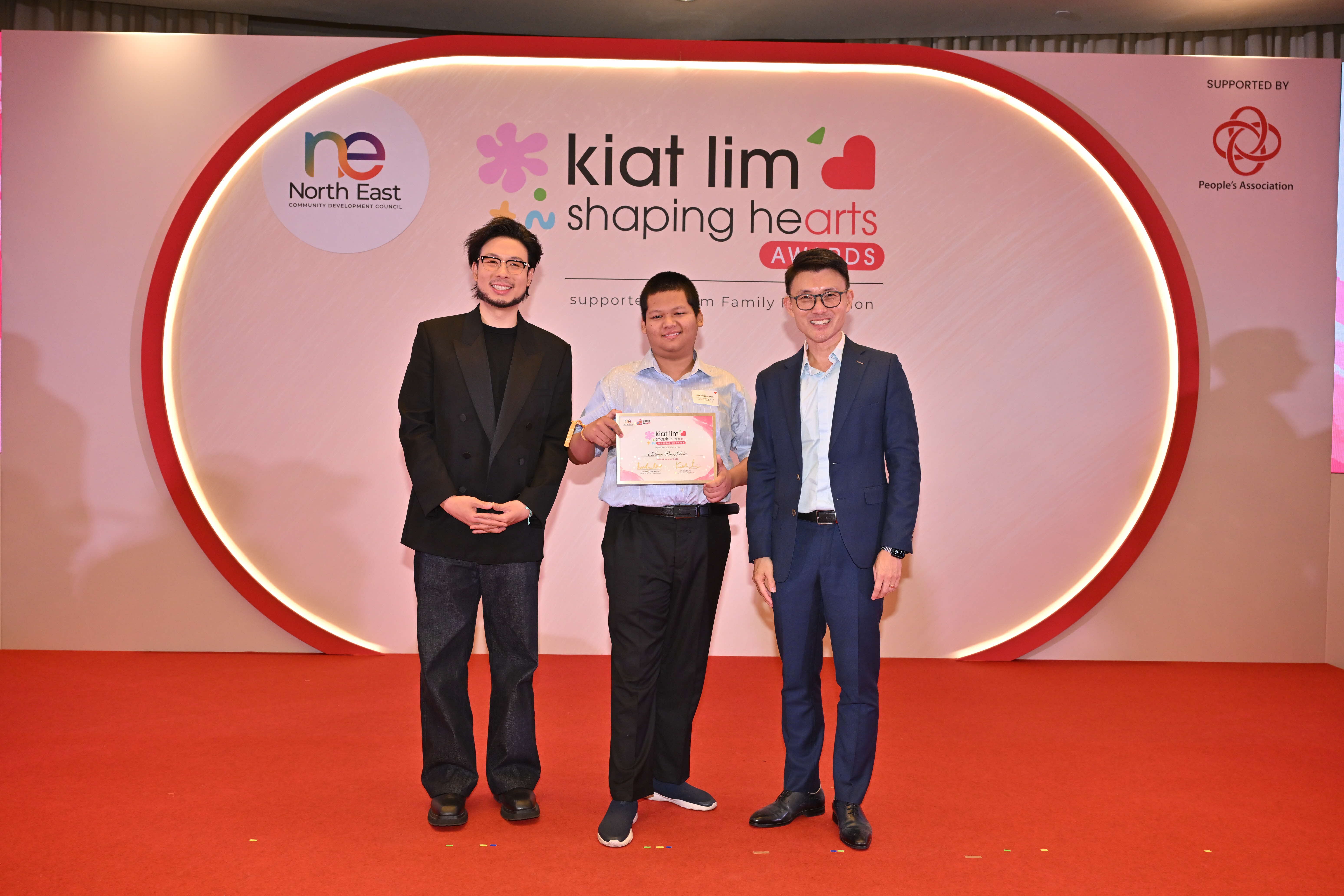 Award recipient standing on stage with the presenter, holding their certificate and posing for a group photo during the award ceremony, with a backdrop reading ‘Kiat Lim-Shaping Hearts Awards'.